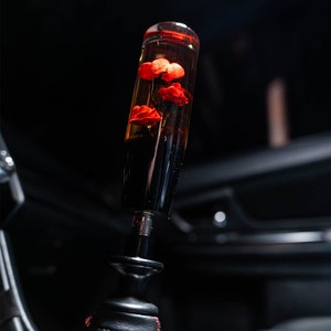 Könnte beinhalten: Ein schwarzer und transparenter Schaltknauf mit roten Blumen, die in Harz eingebettet sind. Der Knauf ist an einem schwarzen Lederschaltmanschetten befestigt.