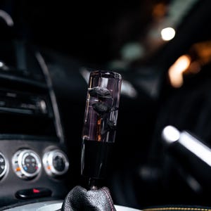 Puede incluir: Un pomo de palanca de cambios de coche cilíndrico transparente con inclusiones florales negras. El pomo está montado sobre una bota de cuero negro con costuras rojas, sobre un interior oscuro. El salpicadero y otros controles del coche son visibles en el fondo.