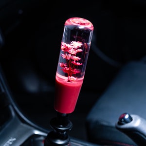 Könnte beinhalten: Ein pinkfarbener und transparenter Schaltknauf mit einem kleinen Strauß rosa Blumen, die in Harz gegossen sind.