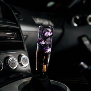 May include: A custom car shift knob with a clear, cylindrical design. The knob is filled with purple flower-shaped elements. The base is gold-colored, and it is mounted on a black shift boot within a car's interior.