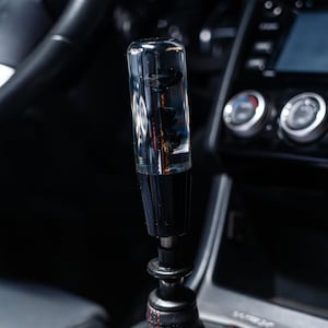 May include: A transparent, cylindrical gear stick knob with a black base and decorative elements inside. The gear stick is in a car, with a black leather gear stick gaiter and red stitching. The car's interior is dark.