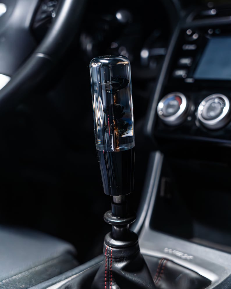 May include: A transparent, cylindrical gear stick knob with a black base and decorative elements inside. The gear stick is in a car, with a black leather gear stick gaiter and red stitching. The car's interior is dark.