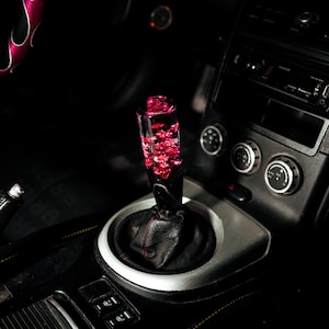 May include: A custom car gear shift knob with a clear, cylindrical design filled with red floral accents. The knob is mounted on a black leather boot and surrounded by a silver trim, set within a dark car interior.