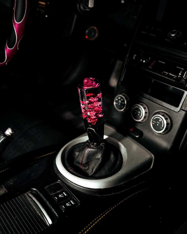 May include: A custom car gear shift knob with a clear, cylindrical design filled with red floral accents. The knob is mounted on a black leather boot and surrounded by a silver trim, set within a dark car interior.