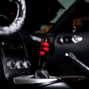 Puede incluir: Primer plano del interior de un coche con un pomo de cambio personalizado. El pomo es transparente y contiene rosas rojas. El interior circundante es negro, incluyendo el salpicadero, la consola central y el volante.