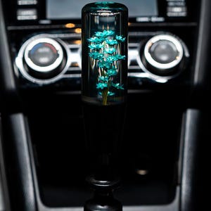 Peut inclure: Un pommeau de levier de vitesse noir avec un cylindre transparent contenant des fleurs bleues à l'intérieur. Le pommeau est installé sur le levier de vitesse d'une voiture.
