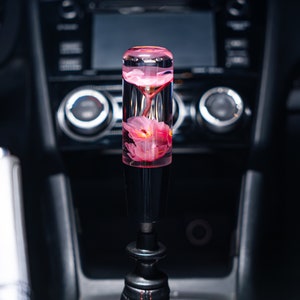 May include: A black gear shift knob with a clear cylinder containing a pink flower inside. The cylinder is filled with a clear liquid.