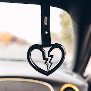 Peut inclure: Un charme de voiture noir en forme de cœur brisé avec un éclair à l'intérieur. Le charme est suspendu au rétroviseur d'une voiture.