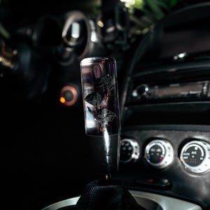 May include: A clear, cylindrical gear stick knob with a black floral design inside. The knob is mounted on a black leather gaiter, set against a dark car interior with gauges and controls.