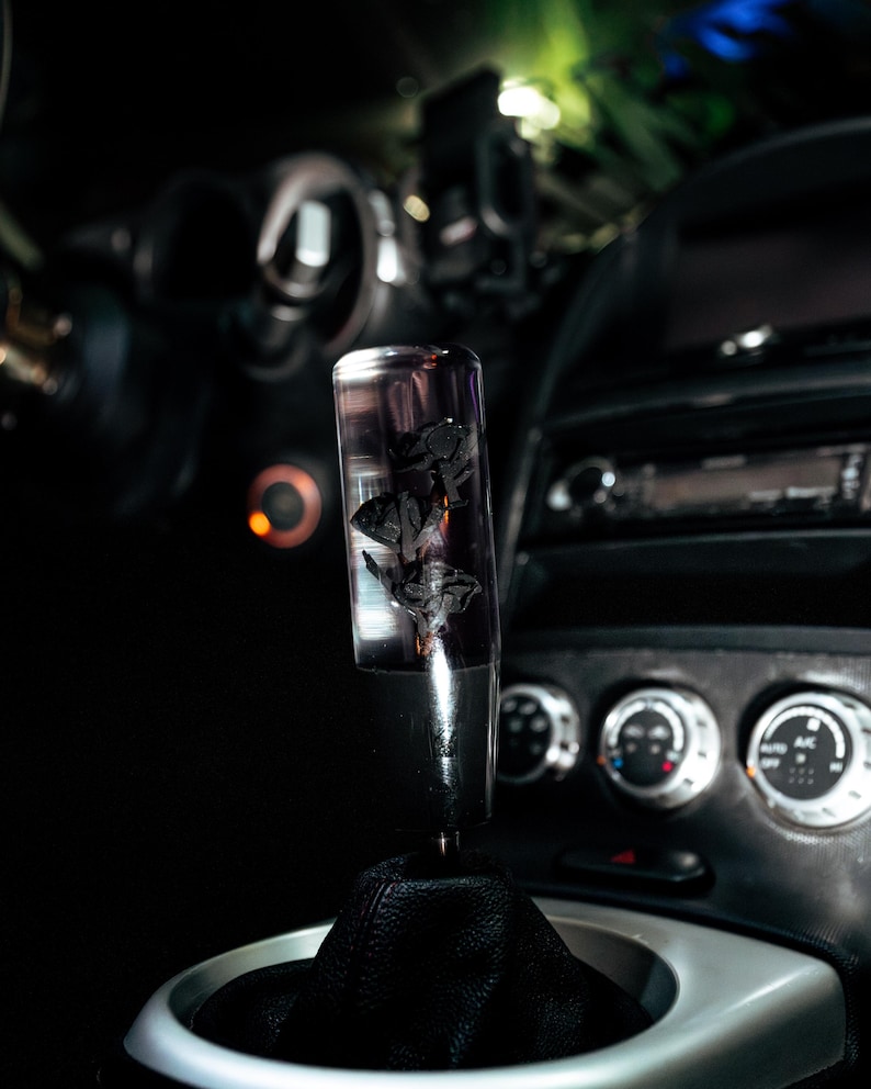 May include: A clear, cylindrical gear stick knob with a black floral design inside. The knob is mounted on a black leather gaiter, set against a dark car interior with gauges and controls.