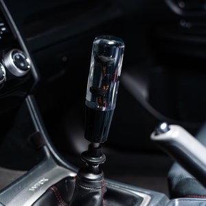 Könnte beinhalten: Ein schwarzer und transparenter, kundenspezifischer Schaltknauf mit einem floralen Design im transparenten Teil. Der Knauf ist an einem schwarzen Lederschaltmanschetten mit roten Nähten befestigt. Die Schaltmanschette ist mit "WRX" beschriftet.