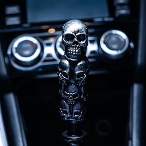 Könnte beinhalten: Ein schwarzer Schaltknauf aus Metall mit drei übereinander gestapelten silbernen Totenköpfen.