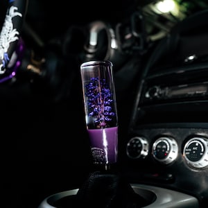Puede incluir: Pomo de palanca de cambios de coche personalizado con un diseño floral púrpura encapsulado en resina transparente. El pomo está montado en una funda de cuero negro, con un ribete plateado. El interior del coche es oscuro.