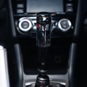 May include: A transparent, cylindrical gear shift knob with a black base and a black leather boot with red stitching. The knob is in a car interior with a dark dashboard and console.