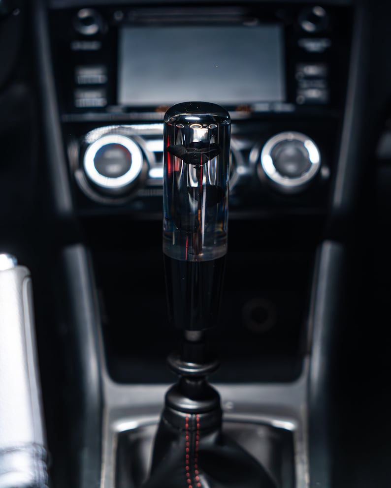 May include: A transparent, cylindrical gear shift knob with a black base and a black leather boot with red stitching. The knob is in a car interior with a dark dashboard and console.