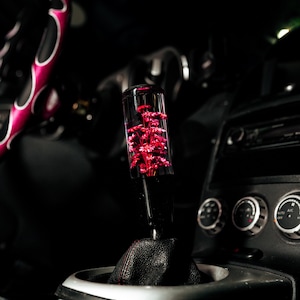 May include: A clear shift knob containing red flowers, mounted on a black leather shift boot. The car interior features a steering wheel with a pink and white design. The background is dark.
