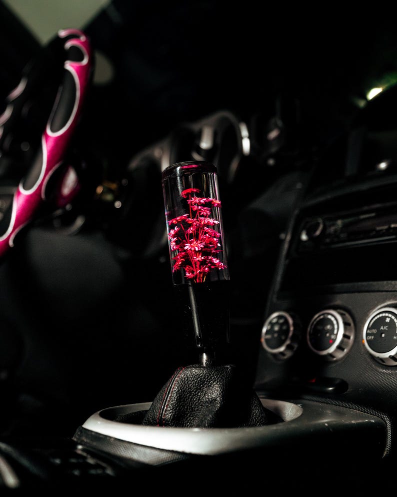 May include: A clear shift knob containing red flowers, mounted on a black leather shift boot. The car interior features a steering wheel with a pink and white design. The background is dark.