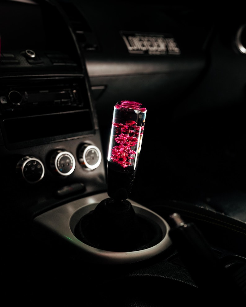 May include: A custom car gear stick with a clear, rectangular body filled with pink floral elements. The gear stick is mounted on a black base within a car's interior, with a silver trim and black dashboard visible in the background.