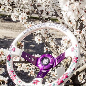 May include: White steering wheel with purple spokes and a pink and white floral design. The steering wheel has the text "Lowered Empire" printed on the top rim.