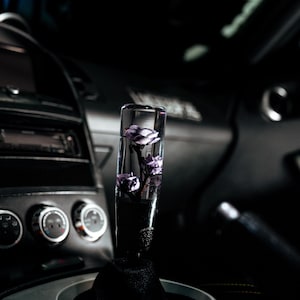 Puede incluir: Un pomo de palanca de cambios de coche cilíndrico transparente con flores moradas en su interior. El pomo se encuentra en un interior oscuro, con el foco en el elemento floral decorativo. La base del pomo es negra y la consola circundante es plateada y negra.