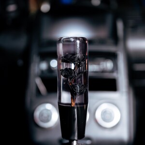 May include: A clear, cylindrical car gear shift knob with black roses encased inside. The bottom portion is solid black, and it is mounted on a black leather shift boot with red stitching. The background is a blurred view of a car's interior.