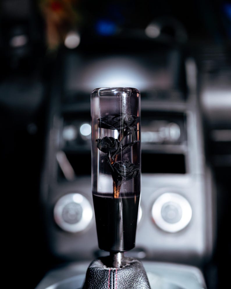 May include: A clear, cylindrical car gear shift knob with black roses encased inside. The bottom portion is solid black, and it is mounted on a black leather shift boot with red stitching. The background is a blurred view of a car's interior.