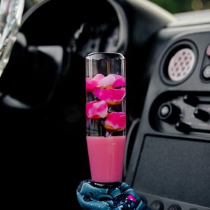 以下が含まれることがあります： ピンク色の液体とピンク色の花びらで満たされた、透明な円筒形の車のギアシフトノブ。 ノブは暗い車の内装に配置され、ベースには模様入りのスリーブが見えます。