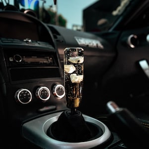 May include: A clear, cylindrical car gear shift knob with white floral accents encased within. The knob is mounted on a black shift boot and is set against a dark interior with silver and black accents. The car's dashboard and controls are visible.