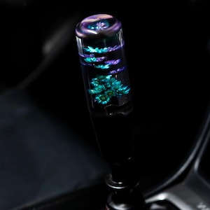 May include: A custom car shift knob with a clear, cylindrical design. The interior features a vibrant display of teal and purple floral elements. The knob is mounted on a black shift lever, set against a dark interior.