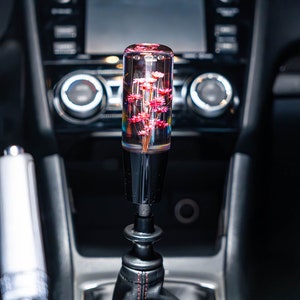 May include: A custom car gear stick knob with a clear, cylindrical design. The knob is filled with pink flowers, creating a unique interior accessory. The base is black, and the gear stick boot is black leather with red stitching.