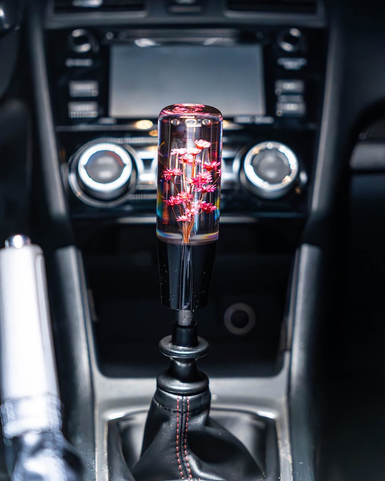 May include: A custom car gear stick knob with a clear, cylindrical design. The knob is filled with pink flowers, creating a unique interior accessory. The base is black, and the gear stick boot is black leather with red stitching.