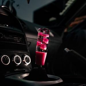 May include: A custom car shift knob with pink rose petals encased in clear resin. The shift knob is cylindrical and mounted on a black base, set against a dark interior. The car's dashboard and controls are visible in the background.