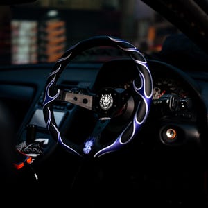May include: Black steering wheel with blue flame design and a silver center. The steering wheel is in a car interior. The center of the wheel has a logo with a skull and the text "LOWSPEED".