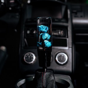 May include: A custom car shift knob featuring three blue roses encased in clear resin. The shift knob is black with a leather boot and red stitching, set against a dark car interior.