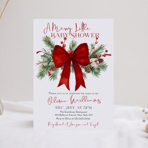 Peut inclure: Une carte d'invitation rectangulaire blanche pour une fête prénatale avec un grand nœud rouge, des branches de pin vertes, des pommes de pin et des baies rouges. Le texte indique A Merry Little BABY SHOWER, Olivia Williams, Dec. 21st. at 5pm, The Grandview Restaurant, 456 Bellevue Avenue, New York, NY, Rsvp to Olivia at 021.000.0456, Registered at Amazon and Target.