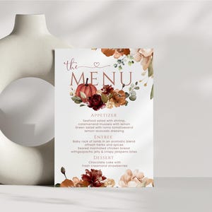 以下が含まれることがあります： 「the MENU」という文字がスクリプト体とセリフ体で記された長方形のメニューカード。カボチャ、濃い赤のバラ、オレンジ色の花の透明水彩画が描かれています。メニューには前菜、メインディッシュ、デザートの項目が記載されています。