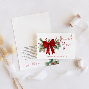 Peut inclure: Une carte de remerciement blanche pli&eacute;e pour une f&ecirc;te pr&eacute;natale avec une illustration aquarelle de branches de pin, de baies rouges et d'un grand n&oelig;ud rouge. Le texte indique Thank You et Olivia's Baby Shower en rouge avec un message imprim&eacute;.