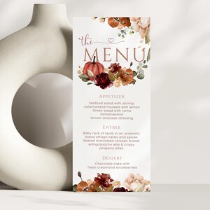 以下が含まれることがあります： 「the MENU」という文字と、カボチャ、濃い赤とオレンジの花、緑の葉の水彩画が描かれた縦型のメニューカード。メニューにはAppetizer、Entree、Dessertのセクションと料理の説明が記載されています。