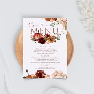 以下が含まれることがあります： 「the MENU」というセリフ体の文字が印刷された長方形のメニューカード。上部と下部にはカボチャ、濃い赤やオレンジの花、緑の葉の透明水彩画が描かれており、背景は白地となっている。