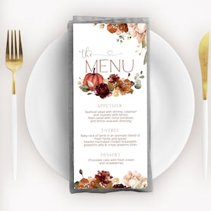 Könnte beinhalten: Eine weiße Menükarte mit floralen und Kürbis-Illustrationen, präsentiert auf einem weißen Teller mit einer goldenen Gabel und einem Messer. Die Speisekarte bietet Vorspeisen, Hauptgerichte und Desserts.