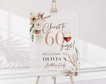 May include: A rectangular birthday party sign featuring the text Cheers to 60 years, Welcome to Olivia's Birthday Party, and 24 June 202X. The design includes a wine glass illustration and floral arrangements in beige, white, and brown tones.