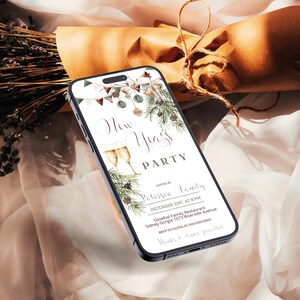 May include: A smartphone displays a New Year's Party invitation with champagne glasses and festive greenery. The phone rests on a soft, draped fabric with dried lavender and a wrapped gift in the background.