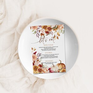 May include: A printable wedding menu card with a fall theme. The card features a watercolor illustration of autumn leaves, flowers, and pumpkins. The menu includes appetizers, entrees, and desserts. The text on the card reads "Let's Eat".