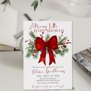 Peut inclure: Une carte d'invitation blanche pour une baby shower orn&eacute;e d'un grand n&oelig;ud rouge, de branches de pin, de pommes de pin et de baies rouges. Le texte indique A Merry Little BABY SHOWER, Olivia Williams, Dec. 21st at 5pm, The Grandview Restaurant.