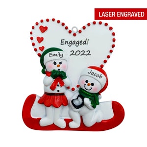 May include: A white heart-shaped ornament with the words "Engaged! 2022" and the names "Emily" and "Jacob" written on it. Two snowmen are depicted on the ornament, one is kneeling and holding a ring box, the other is standing with a surprised expression.