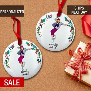 May include: Two white round ceramic ornaments with red ribbon hangers. Each ornament features a colorful illustration of a person dancing with a floral wreath around the image. The text "Emily 2021" is printed on each ornament.