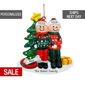 May include: Personalized Christmas ornament featuring a couple in winter attire standing in front of a green Christmas tree with a star on top. The ornament also includes a dog, a red sled, and a yellow gift box. The text "2023", "Nancy", "Nick", "Rex", "Baby", and "The Baker Family" is included on the ornament.