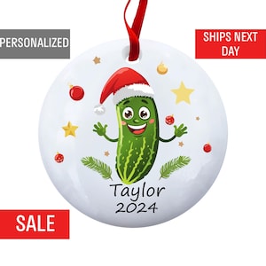 May include: A white round Christmas ornament with a green pickle wearing a red Santa hat. The pickle is smiling and has arms and legs. The ornament also has red and gold stars and the text "Taylor 2024".