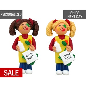 May include: Two personalized Christmas ornaments of girls dressed as artists. The girl on the left has brown hair and is holding a paintbrush and a bottle of glue. Her ornament has the name "Kayla 2022" written on it. The girl on the right has blonde hair and is holding a paintbrush and a bottle of glue. Her ornament has the name "Chloe 2022" written on it.