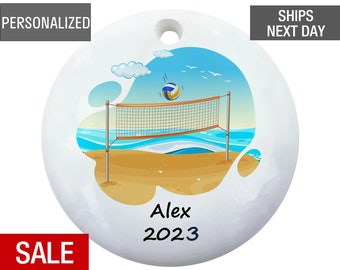 May include: White round ceramic ornament with a beach volleyball scene. The ornament features a volleyball net, a volleyball, a beach, and blue sky with clouds. The text "Alex 2023" is written on the ornament.